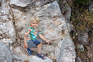 Child, blond toddler boy, standing on a big rock in the mountains, smiling