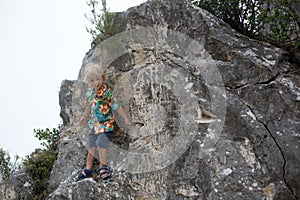 Child, blond toddler boy, standing on a big rock in the mountains, smiling