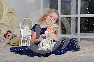 Child blonde in a blue dress sitting on the bed with a flashlight and a Russian New Year`s doll in a kokoshnik