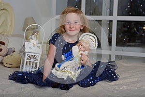 Child blonde in a blue dress sitting on the bed with a flashlight and a Russian New Year`s doll in a kokoshnik