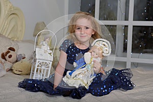 Child blonde in a blue dress sitting on the bed with a flashlight and a Russian New Year`s doll in a kokoshnik