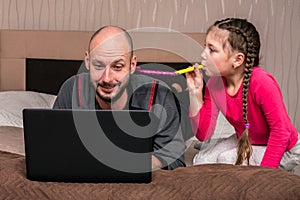 A child blows a festive pipe near the man`s ear. A little girl prevents dad from working on his laptop in the bedroom. Family is