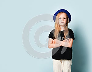 Child in blue hat, black t-shirt and white pants. She is looking at you with crossed hands while posing against turquoise studio
