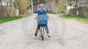 Child in blue vest and black leggings rides balance bike on dirt road, in countryside