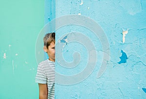 Child by blue the wall, baby fashion concept. cute boy in the summer on the street. hid around the corner, looks out and laughs