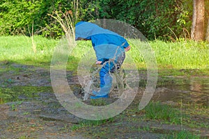 A child in a blue waterproof overalls is jumping into a puddle in nature. Splashing water. Summer, sunny day. The idea