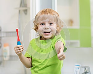 Child boy brushing teeth in bath and showing thumbs up.