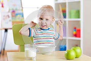 Child boy eating healthy food and showing his strength indoors