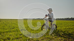 Child boy in hat is riding a bike with pinwheel on green field in country road.