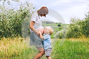 Child boy kissing belly of pregnant her mother on nature backgro