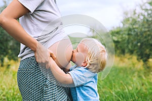 Child boy kissing belly of pregnant her mother on nature backgro