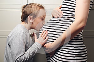 Child boy kissing his mother pregnant tummy. Pregnant mother and son hugging. Cute boy with mom spending time together at home