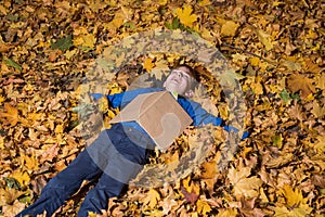 Child boy lies in fallen autumn leaves with the book on his chest. Boy resting in fall forest