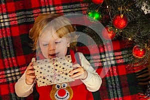 Child boy lies on a plaid near the christmas tree and examines a gift box