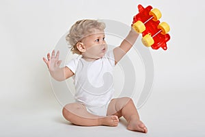 Child boy playing with toys indoors, sitting on floor and raising hands up, holding red and yellow toy car, looking at it with big