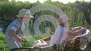 Child boy pushing wheelbarrow with little girl at village, children in straw hats rest at lake