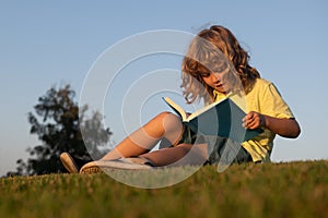 Child boy reading book, laying on grass in field on sky background. Portrait of clever kids. Kids success, successful