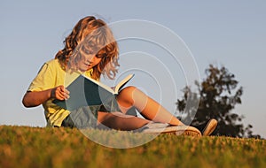 Child boy reading book, laying on grass in field on sky background. Portrait of clever kids. Kids success, successful