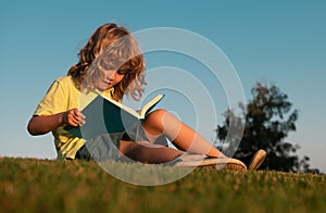 Child boy reading book, laying on grass in field on sky background. Portrait of clever kids. Kids success, successful