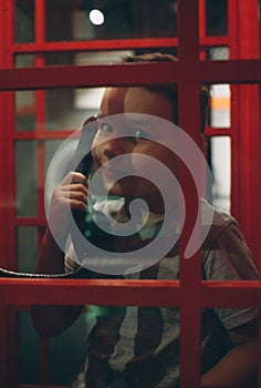 Child boy in a red phone booth smiles and looks at the camera through the window. The boy holds in his hand the receiver