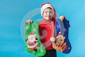 Child boy in red sweater and Santa hat with Christmas socks on blue background with space for text