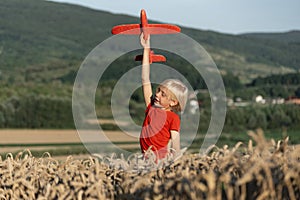 Child boy in red T-shirt plays with toy plane in wheat field on mountains background. Concept of flights and travel with children