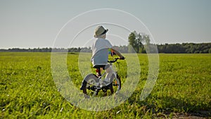 Child boy is riding bike with pinwheel on green field in countryside, back view.