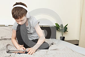 Child boy sitting on bed with pc tablet with wireless headphones