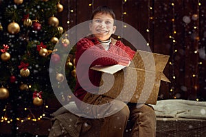 Child boy sitting with gift box near christmas decorated fir tree, dark wooden background