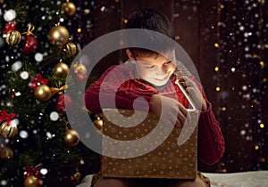 Child boy sitting with gift box near christmas decorated fir tree, dark wooden background