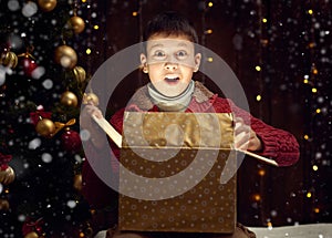 Child boy sitting with gift box near christmas decorated fir tree, dark wooden background