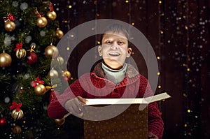 Child boy sitting with gift box near christmas decorated fir tree, dark wooden background