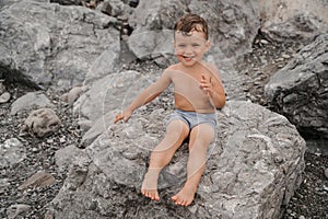 The child boy is sitting, smiling and sunbathing on large boulders, on the beach near the sea.