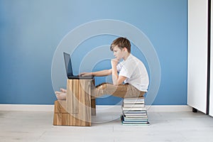 Child boy sitting on stack of books typing on his laptop, studying online at home, modern interior with blue wall background