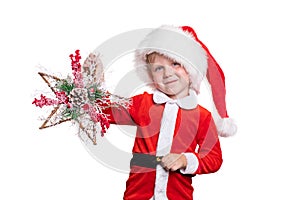 A child boy in a suit and a Santa Claus hat holds a wooden star decoration with berries branches and a pine cone in his hands. New