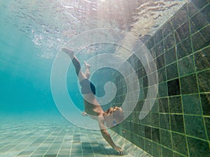 A child boy is swimming underwater in a pool, smiling and holding breath, with swimming glasses