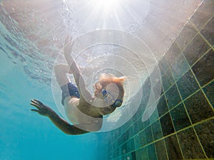 A child boy is swimming underwater in a pool, smiling and holding breath, with swimming glasses