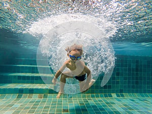 A child boy is swimming underwater in a pool, smiling and holding breath, with swimming glasses
