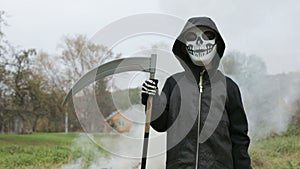 Child boy wearing in scary Halloween skull skeleton mask holding up a scythe in hand