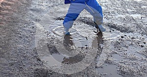 Child in bright blue overalls and rubber boots walk and jump in muddy puddle of black water and earth. Concept about