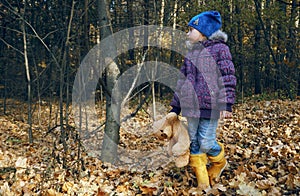 A child in bright rubber yellow boots stands and holds a teddy bear`s paw in his hand. Stuffed toy, best friend. Autumn, dry