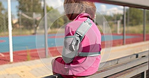 A child with a broken arm outdoors near a playground fence.