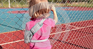 A child with a broken arm outdoors near a playground fence.