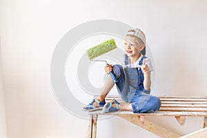 A child Builder sits on a construction ladder in an apartment with white walls and a roller in his hands and shows a thumbs up, a