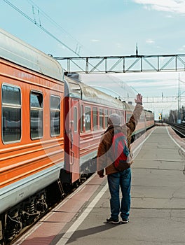 A child bundled up in winter clothing extends a hand high to wave at a departing orange train on a clear day. photo