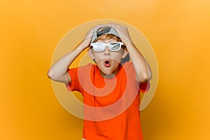 Child in a cap and glasses for watching movies grabbed his head with both hands and twisted his lips into a tube and shows