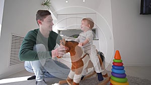 Child care, loving papa playing with merry toddler boy on plush steed seating and swinging at home in kitchen