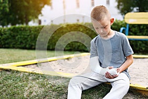 child caucasian with broken limb outdoors sits near the playground and looking on the plaster bandage on his arm. the