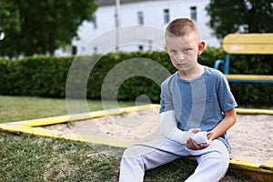 child caucasian with broken limb outdoors sits near the playground and looking on the plaster bandage on his arm. the