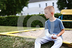 child caucasian with broken limb outdoors sits near the playground and looking on the plaster bandage on his arm. the